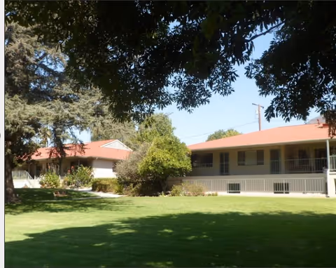 Two single-story buildings with red roofs facing a shaded grassy courtyard with large trees.
