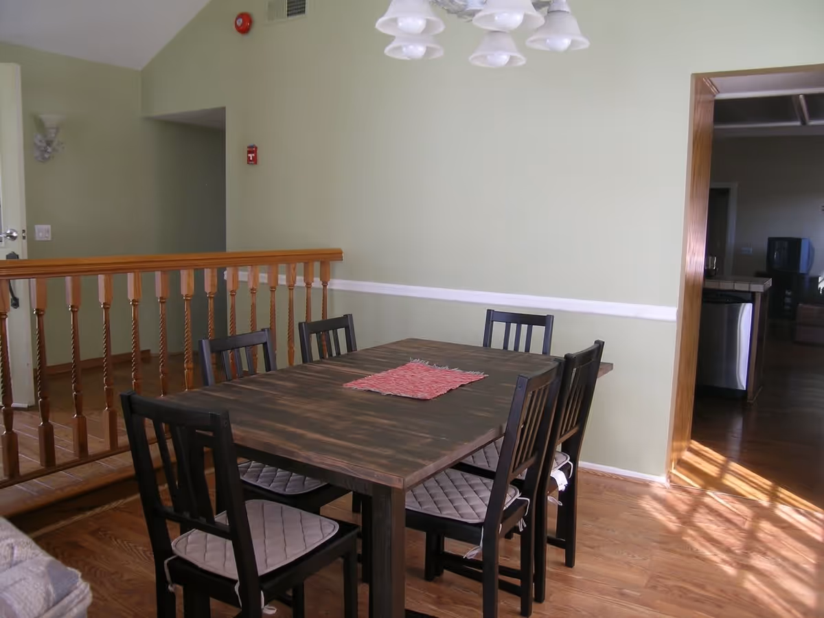 A dining area with a dark wooden table and six black chairs with beige cushions. The walls are painted light green with a white chair rail. There is a wooden railing on the left side and an open doorway on the right leading to another room with a kitchen visible. A ceiling light fixture with multiple white lampshades hangs above the table.