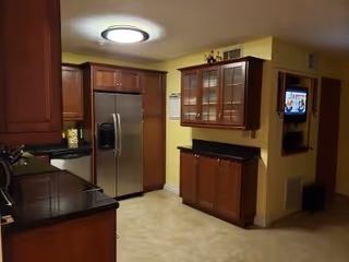 Interior view of a kitchen with wooden cabinets, a stainless steel refrigerator, black countertops, beige tiled floor, and a wall-mounted TV in the adjacent room.