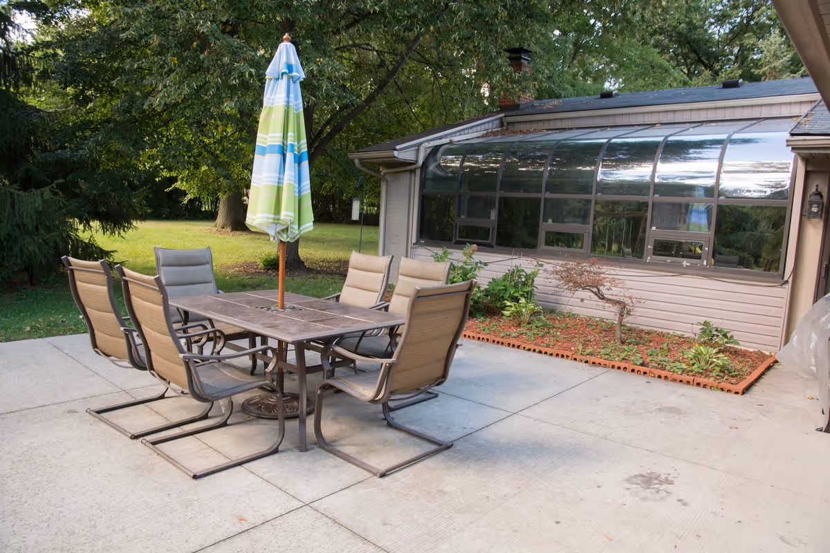 Outdoor patio area with a rectangular table surrounded by six cushioned chairs. A closed umbrella with green, blue, and white stripes is positioned in the center of the table. The patio is adjacent to a building with a large curved glass window and a small garden bed with plants and mulch. Trees and grass are visible in the background.