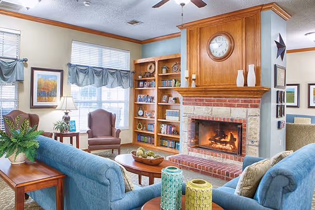 A cozy living room with a lit brick fireplace, wooden mantel with a clock and decorative vases, blue upholstered sofas, a wooden coffee table with decorative items, a side table with a lamp and plant, a brown armchair near windows with blue valances, and a built-in wooden bookshelf filled with books and decor.