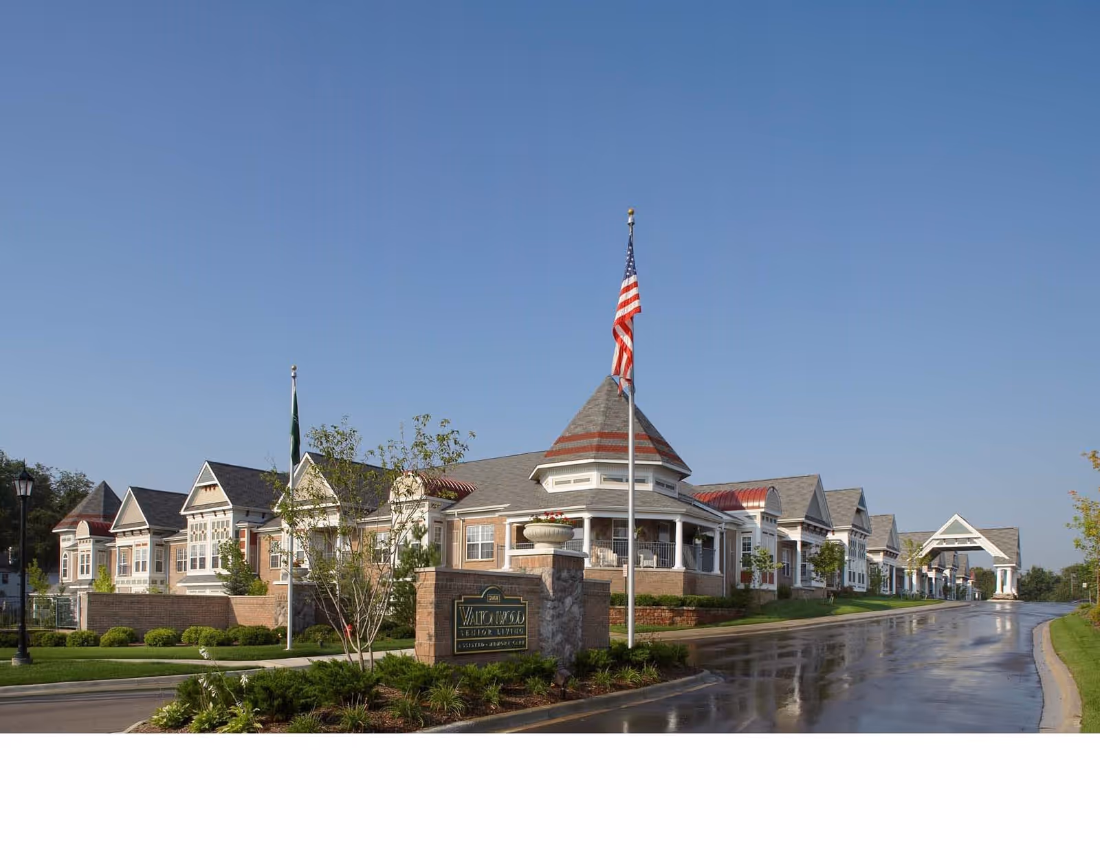 Front exterior of Waltonwood Main senior living building with flagpoles, a covered entrance drive, and landscaped grounds under a clear blue sky.
