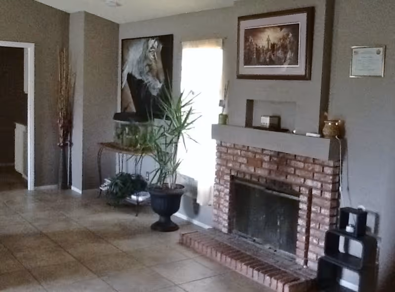 Interior view of a room with a brick fireplace, a potted plant, a table with a fish tank, wall art including a tiger portrait, and a window with a curtain. The floor is tiled and the walls are painted gray.