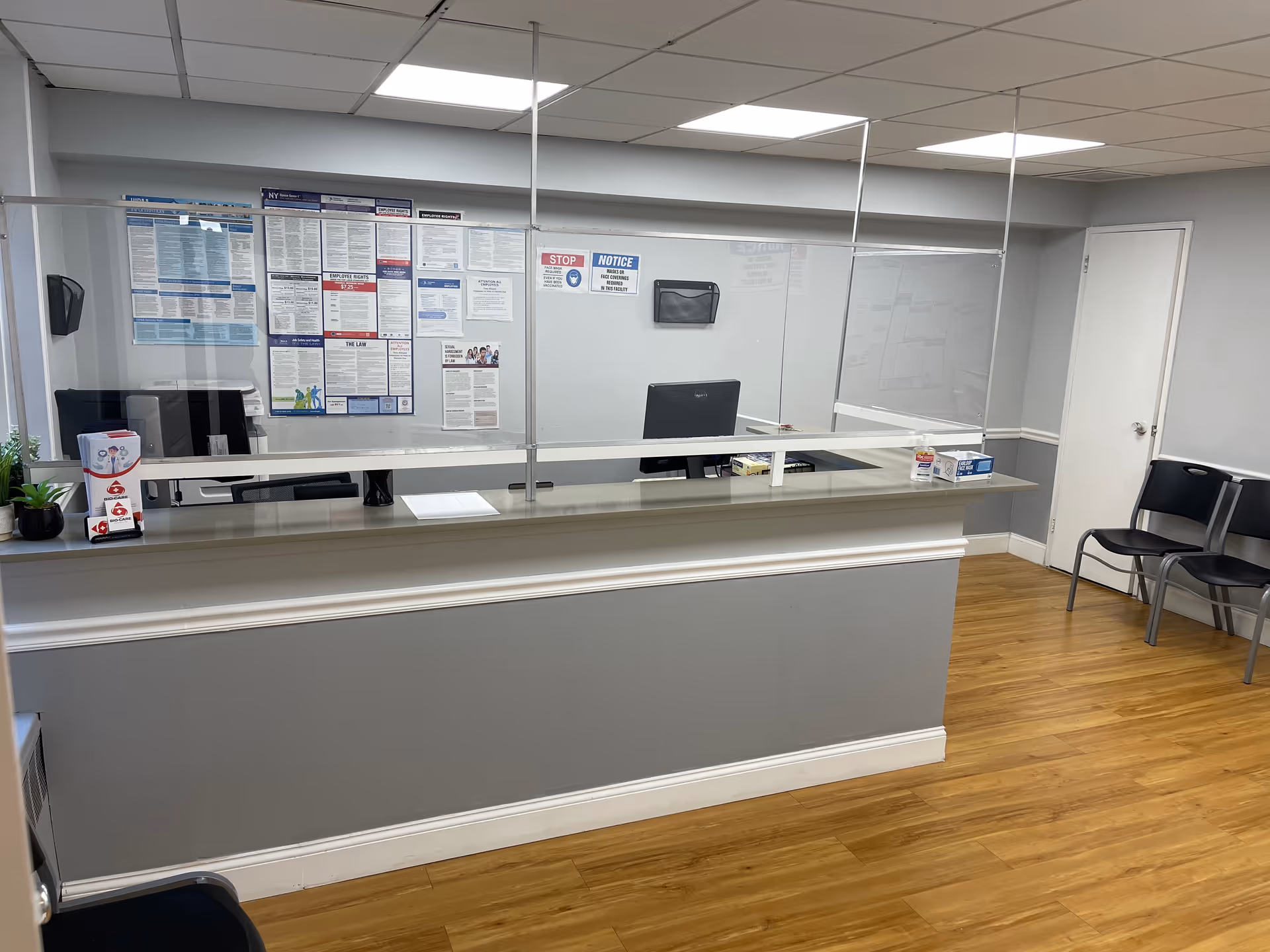 Reception desk with clear dividers, bulletin boards, computer, and empty chairs in a gray interior.