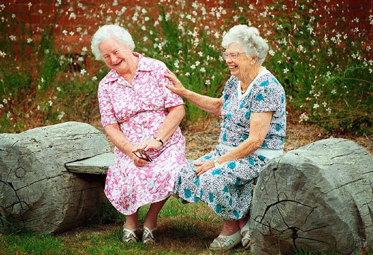 Two elderly women sitting on a rustic wooden bench made from large logs in an outdoor garden area. They are smiling and laughing, enjoying each other's company. One woman is wearing a pink floral dress and holding glasses, while the other is wearing a blue and white patterned dress. The background features greenery and small white flowers.
