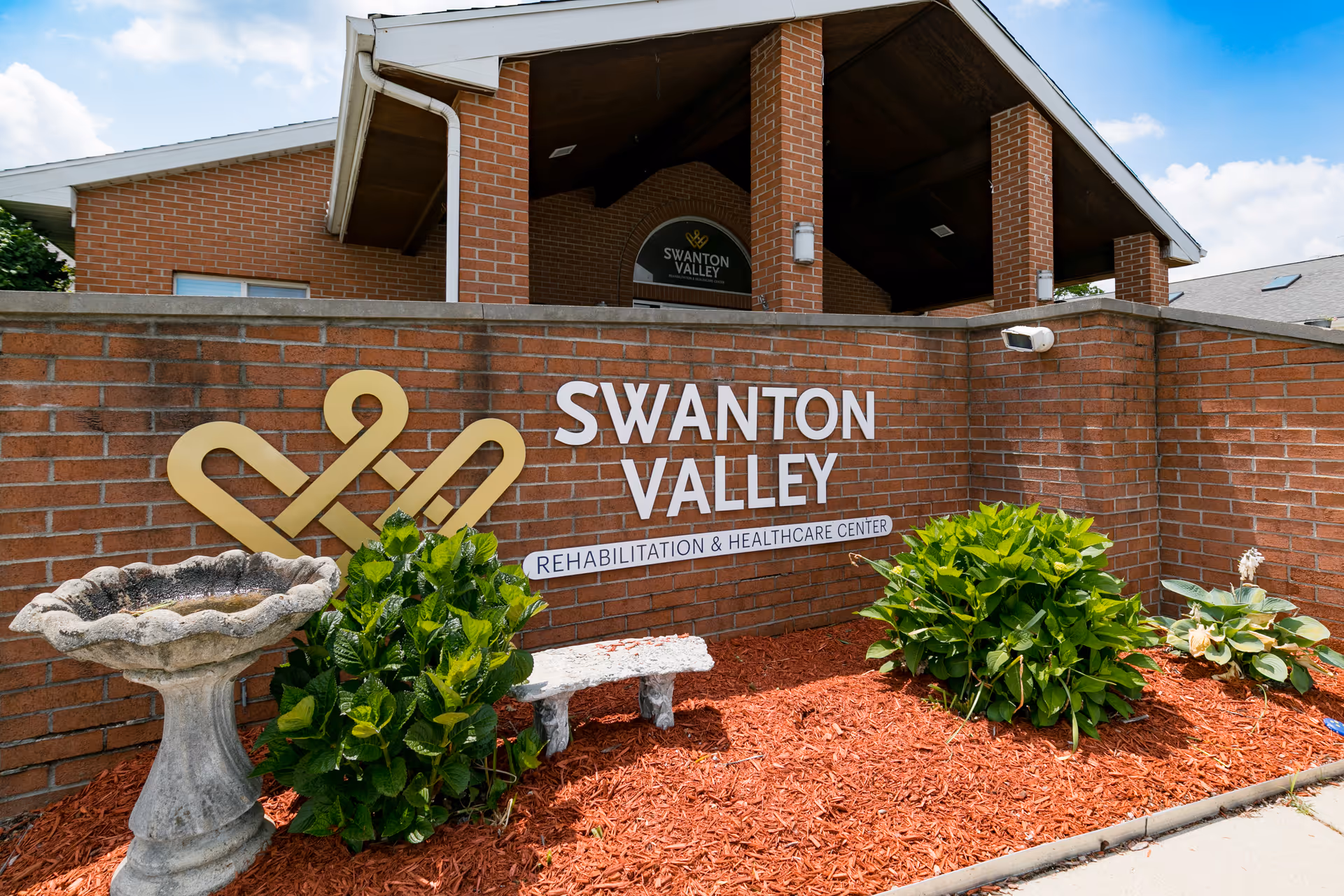 Exterior view of Swanton Valley Rehabilitation and Healthcare Center showing a brick wall with the facility's name and logo, surrounded by landscaping with green bushes, a stone birdbath, and a small stone bench under a partly cloudy sky.