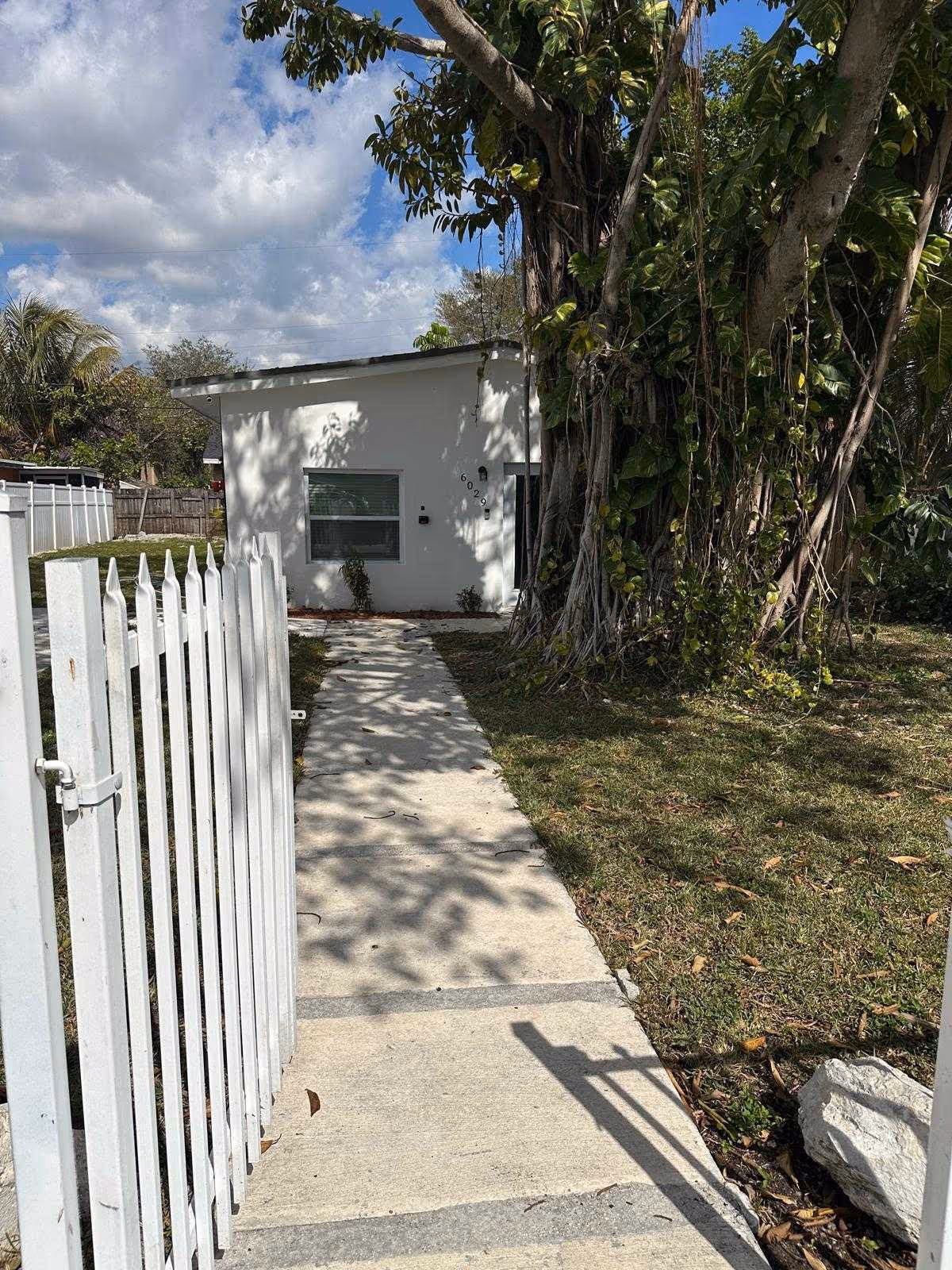 Concrete path leads to a white single-story building, flanked by a picket fence and large tree.