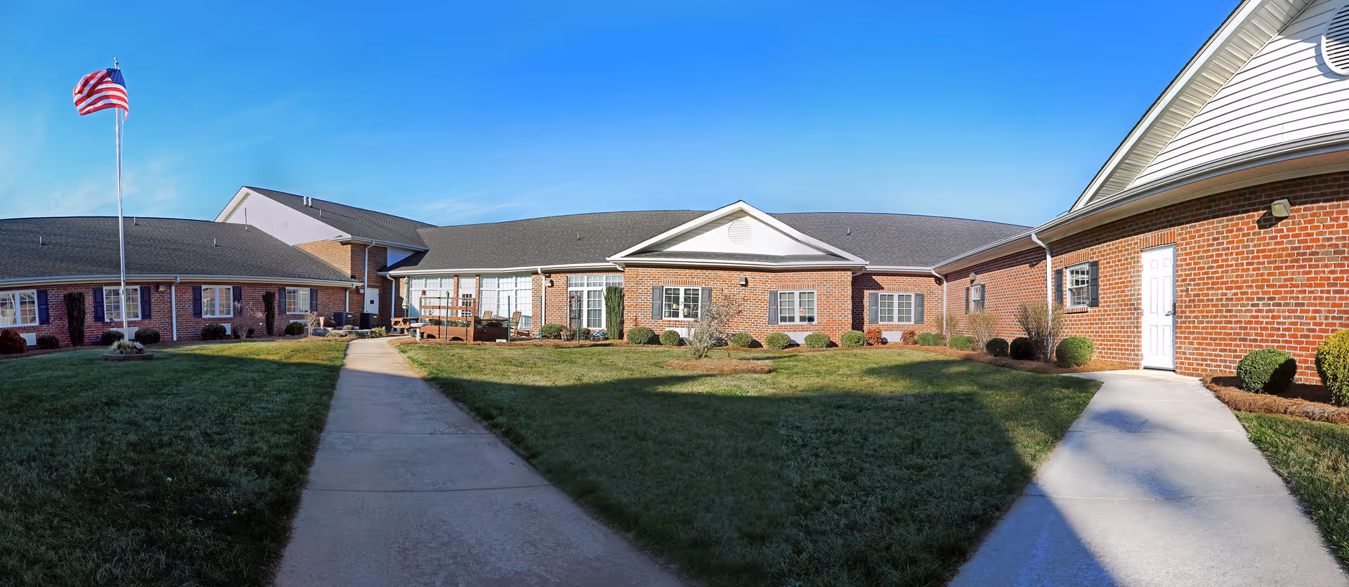 Wide exterior view of a single-story brick building with multiple windows and white doors, surrounded by a well-maintained lawn and a concrete walkway leading through the center. An American flag on a flagpole is visible on the left side under a clear blue sky.