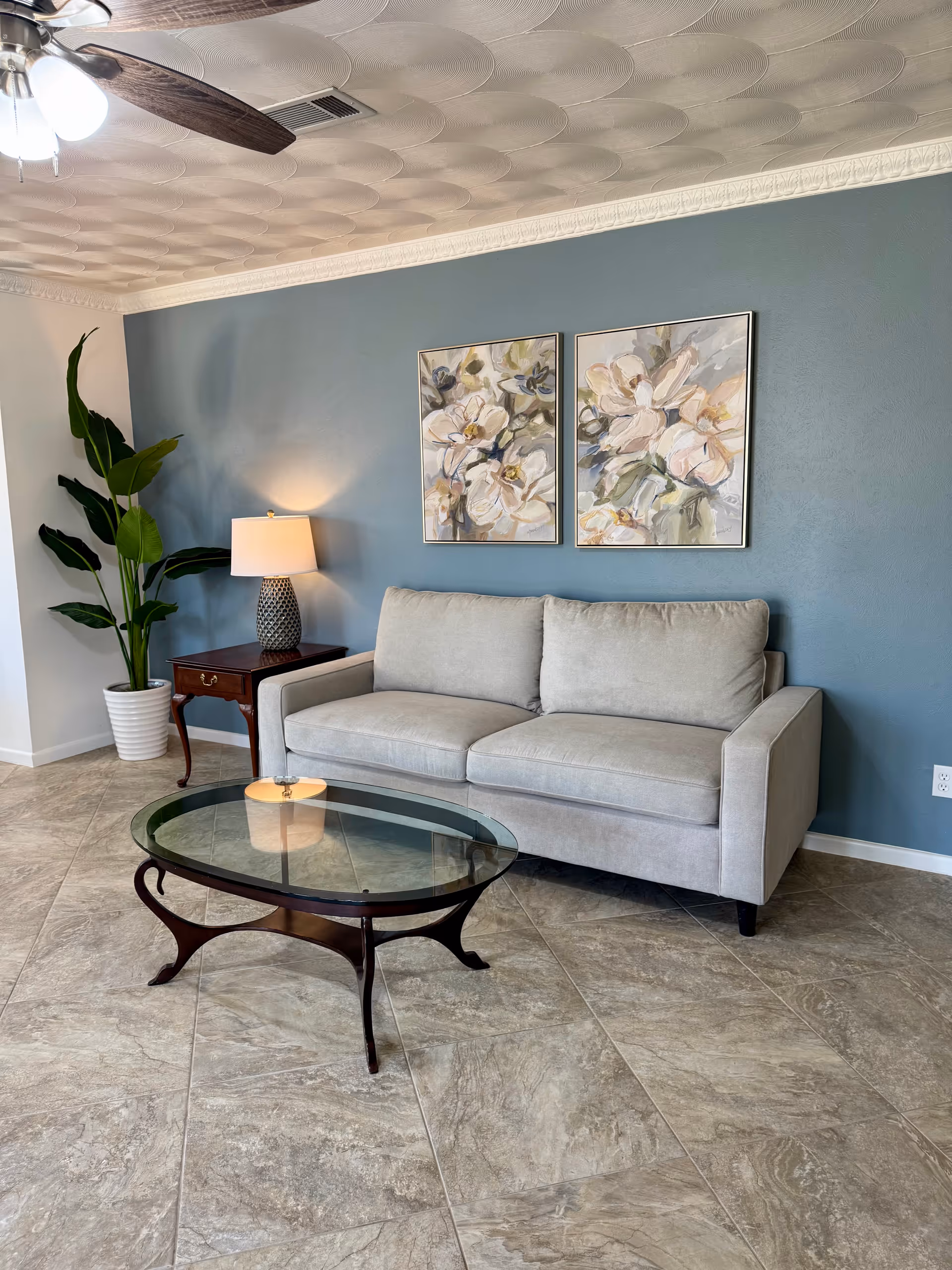 Blue-walled living room with gray sofa, glass coffee table, plant, lamp, and floral art.