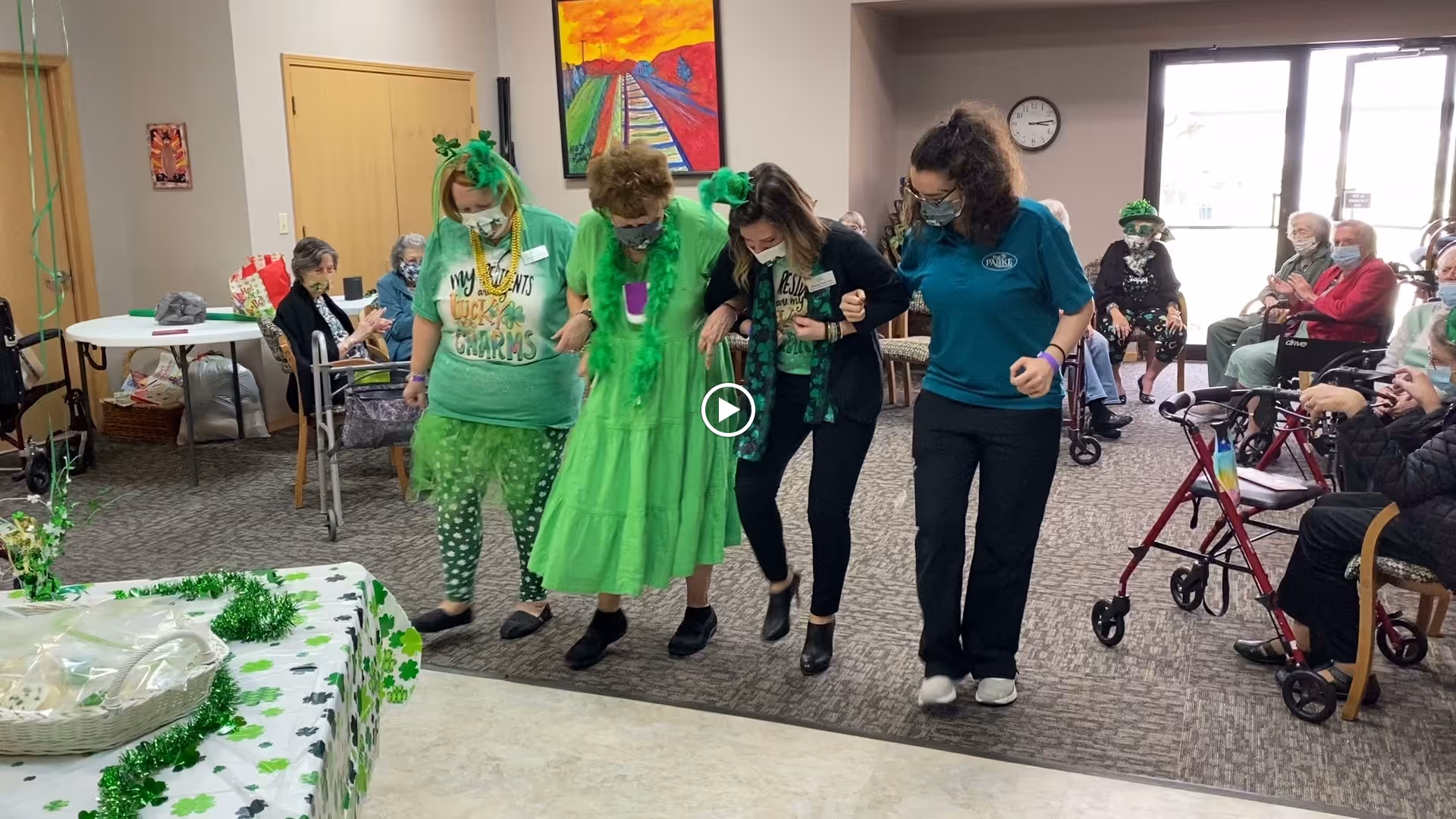 A group of four women, three wearing green festive clothing and accessories, are linked arm-in-arm and dancing or stepping together in a common room of an assisted living facility. Several elderly residents sit around the room watching and clapping, some using walkers or wheelchairs. The room is decorated with green shamrock-themed decorations, suggesting a St. Patrick's Day celebration.