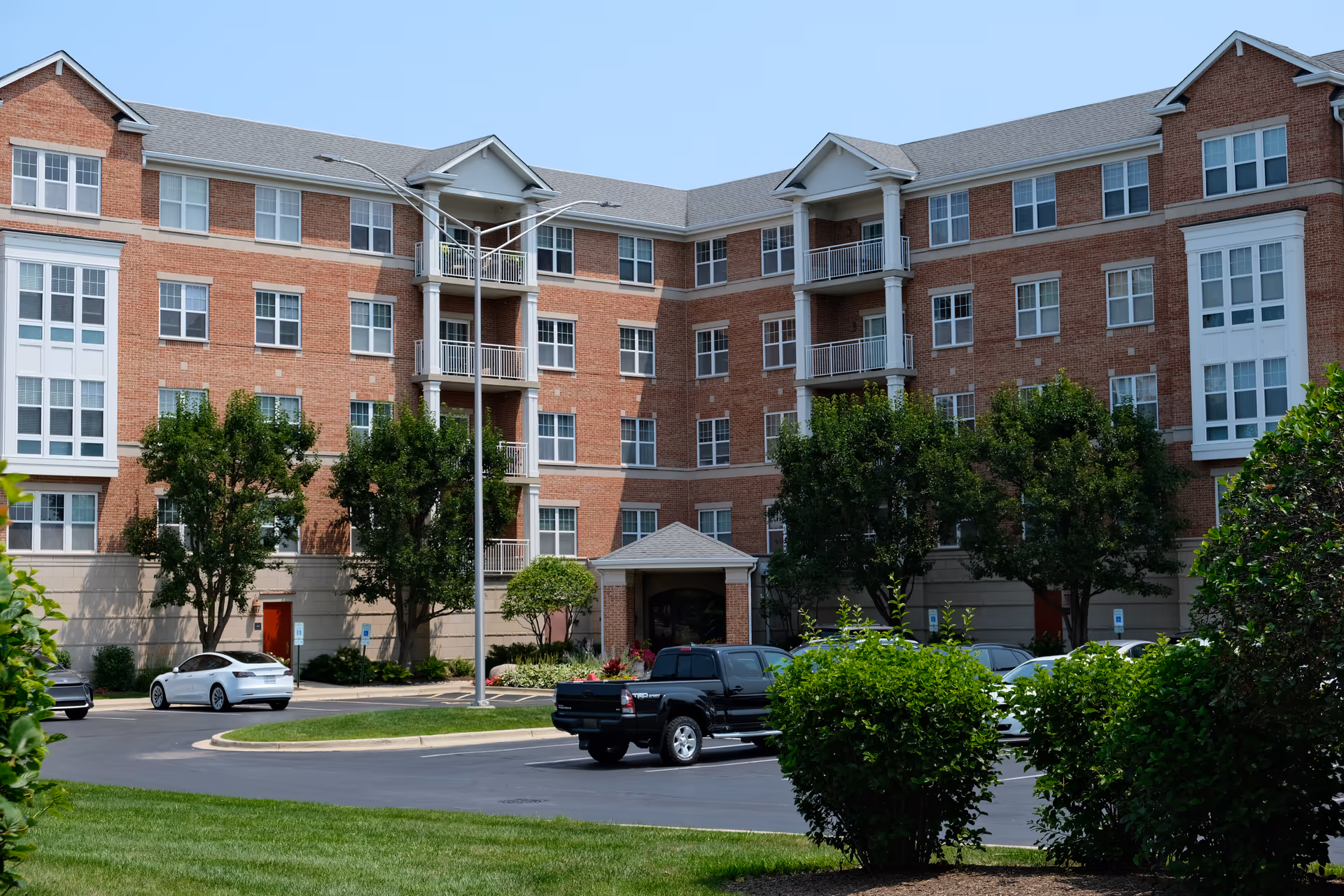 Front exterior of a multi-story brick memory care building with parked cars, trees, and landscaped shrubs.