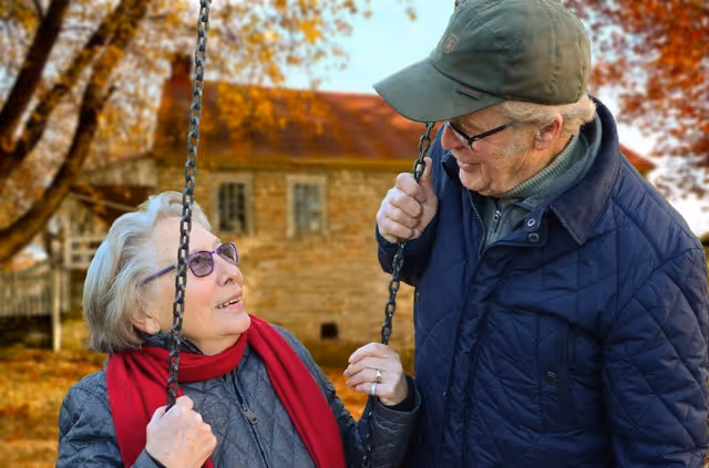 Two elderly people swing outdoors, smiling at each other with autumn trees.