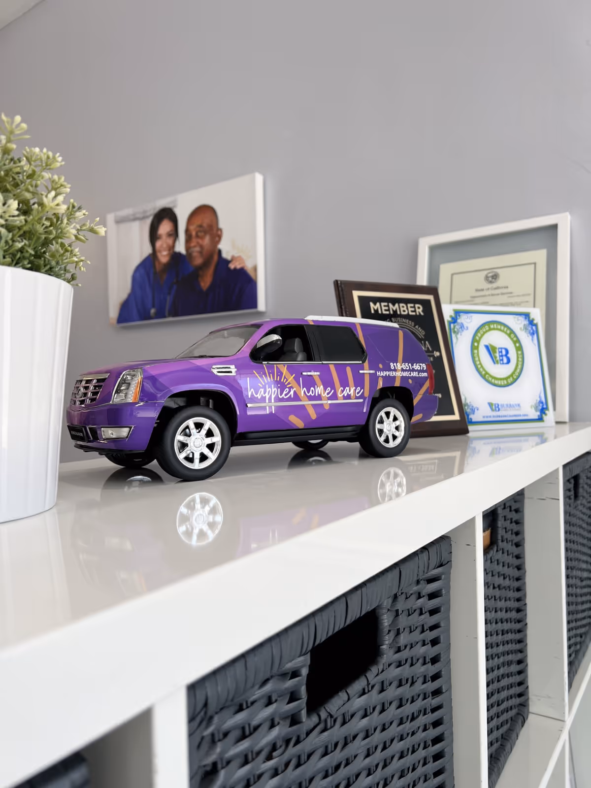 Purple Happier Home Care miniature van sits on a white shelf beside awards.