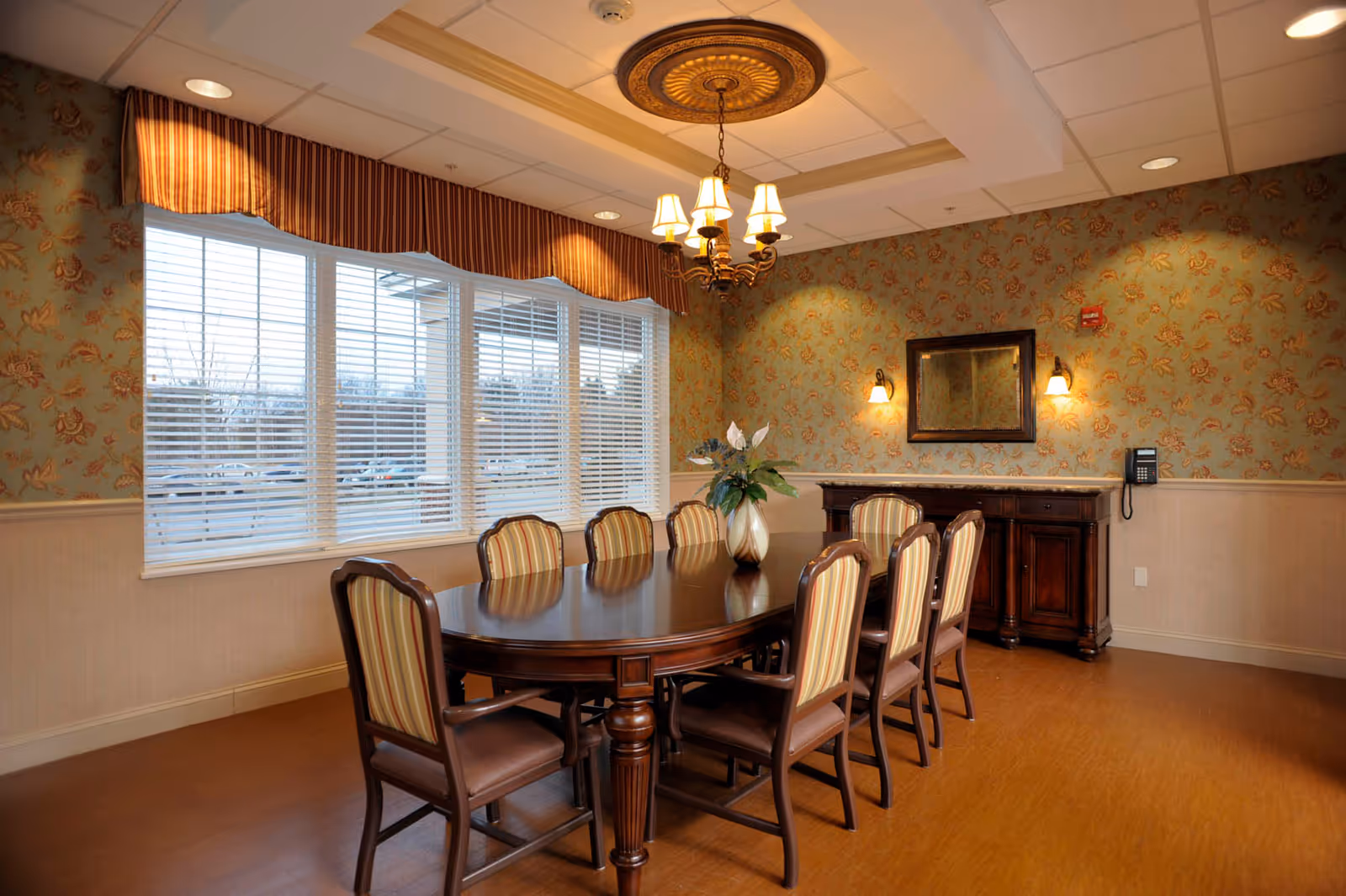A dining room with a large wooden table surrounded by eight striped upholstered chairs. A decorative vase with flowers is placed on the table. The room features floral wallpaper, a wooden sideboard with a mirror above it, wall sconces, and a chandelier hanging from the ceiling. Large windows with white blinds and a striped valance allow natural light into the room.