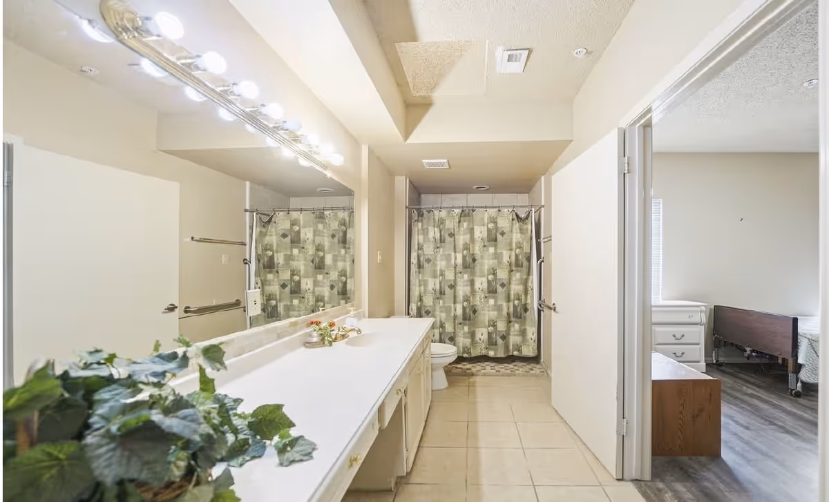 A bathroom with a long white countertop and sink on the left, a large mirror above the countertop with multiple light bulbs, a shower with a patterned curtain at the back, and a toilet next to the shower. To the right, an open door leads to a bedroom with a bed and a white dresser.