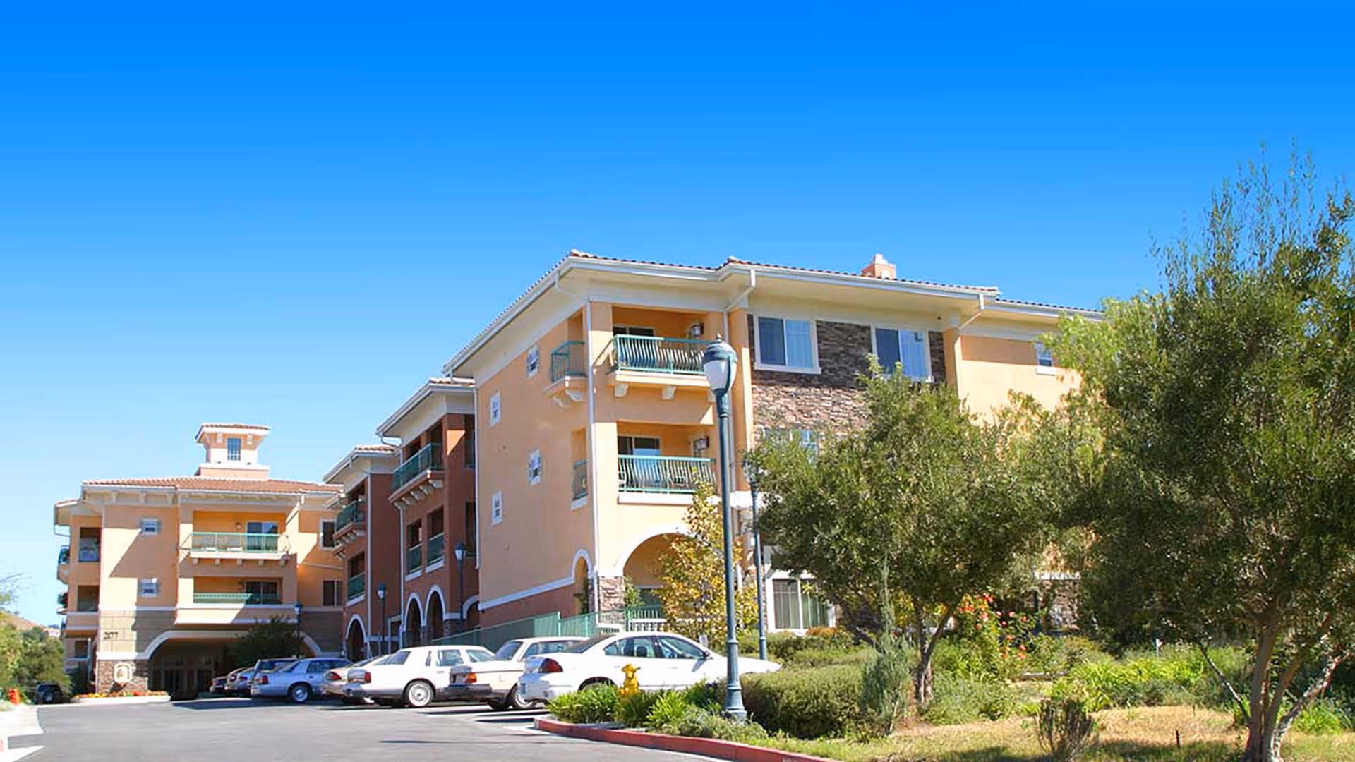 Front exterior of a multi-story Mediterranean-style senior living building with balconies, parked cars, and landscaped trees under a clear blue sky.