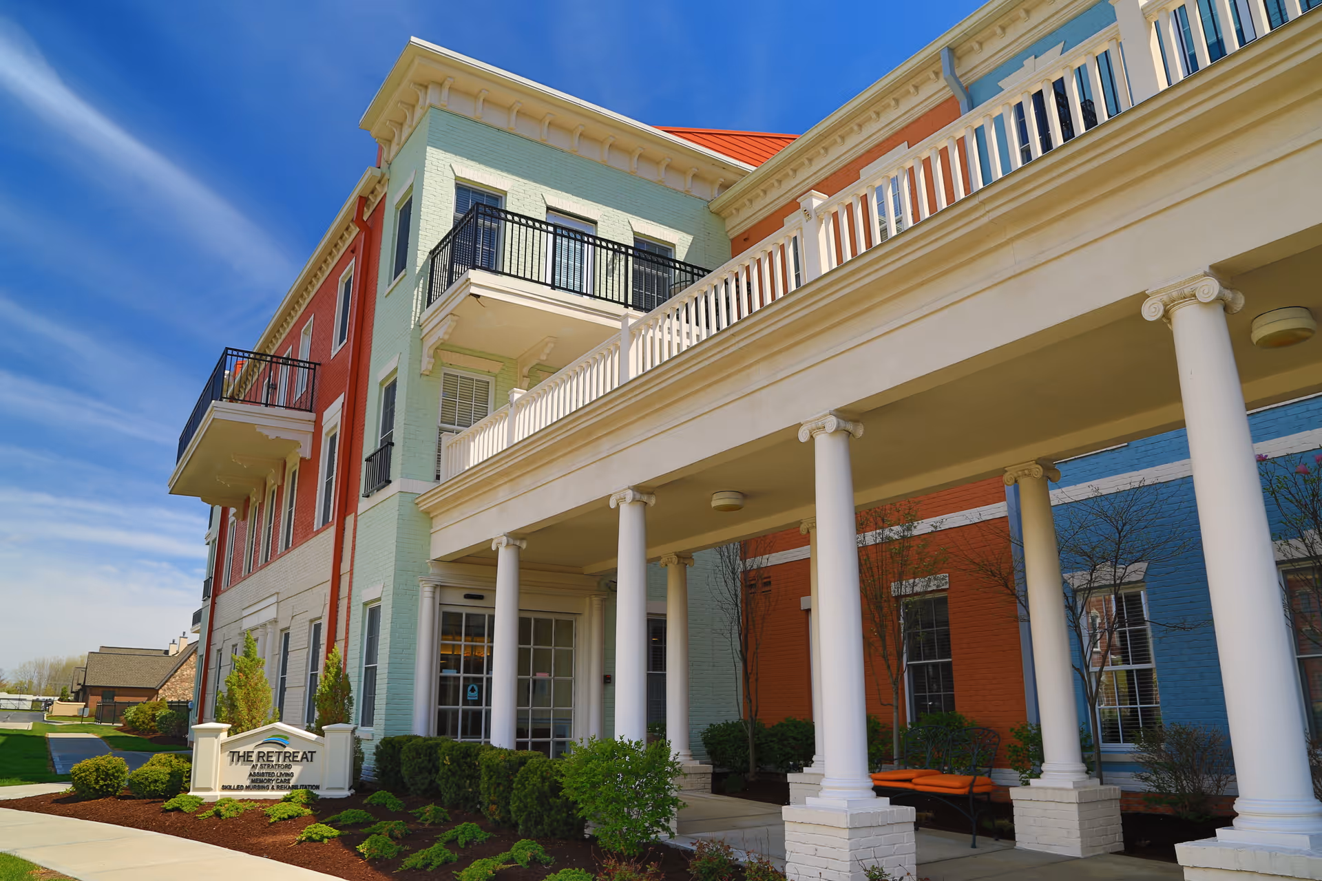 Exterior view of a senior living facility named The Retreat at Stratford, featuring a multi-story building with colorful brick walls in green, red, and blue, white columns supporting a balcony, and landscaped bushes and trees under a clear blue sky.