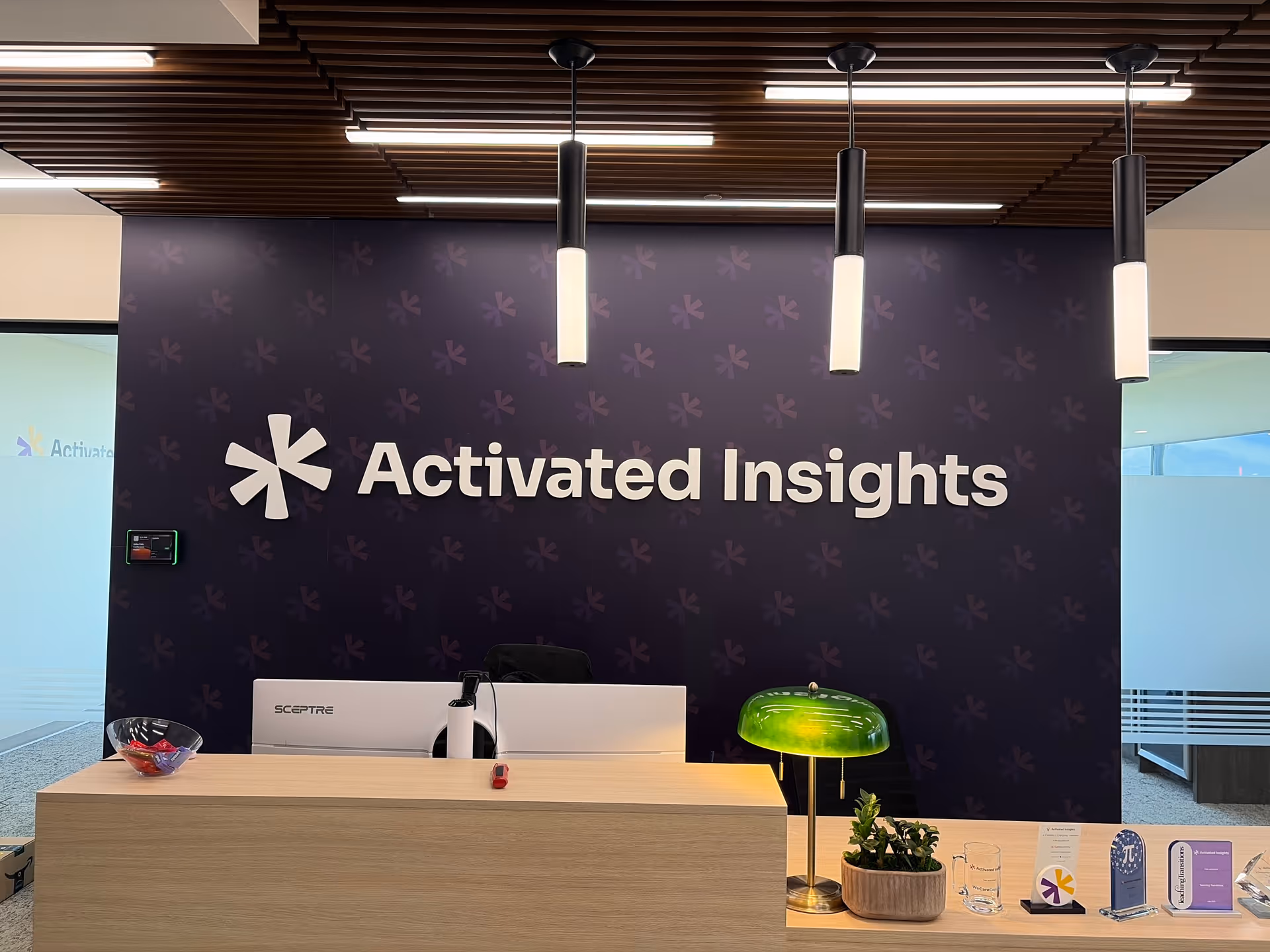 Reception area with wooden desk, dark wall displaying Activated Insights logo, pendant lights, and plants.
