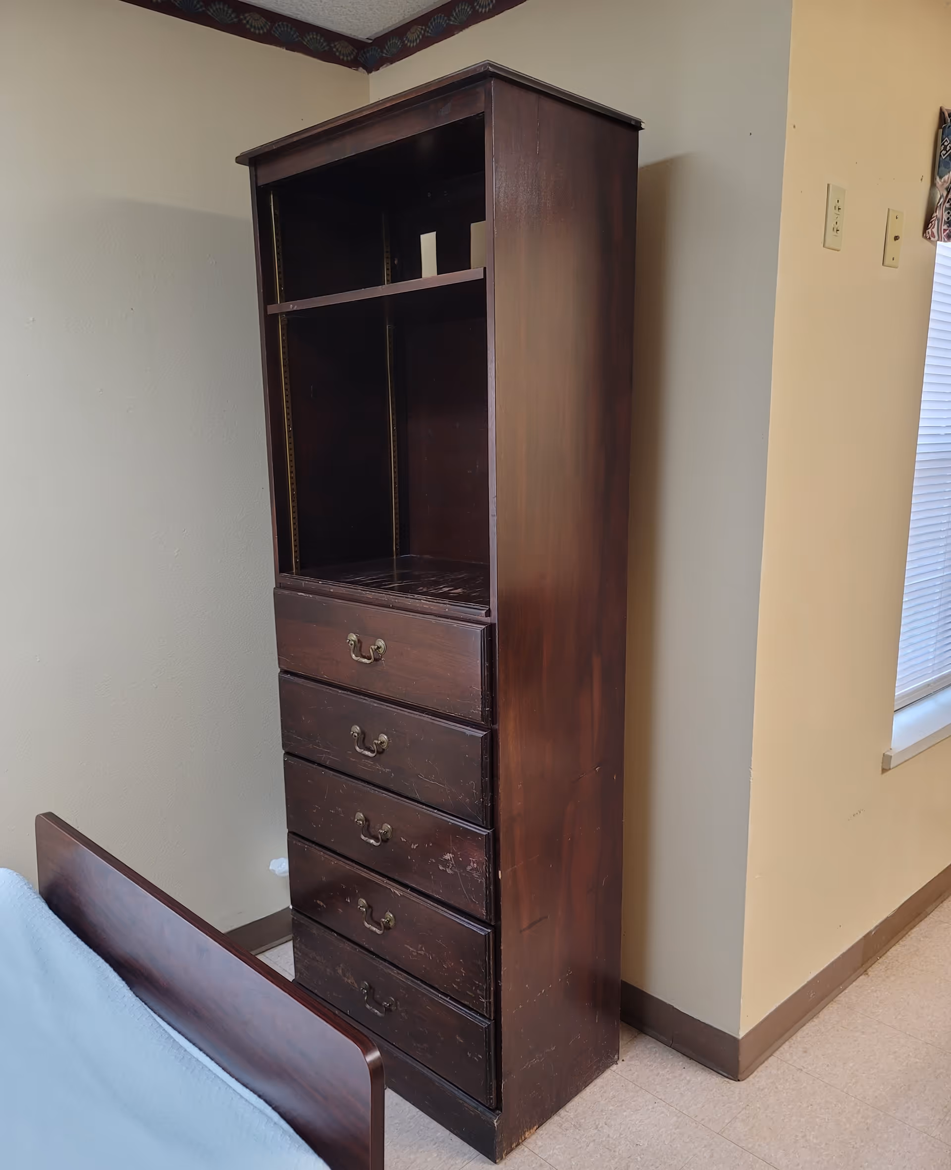 A tall dark wood dresser with drawers stands in a bedroom corner beside a bed.