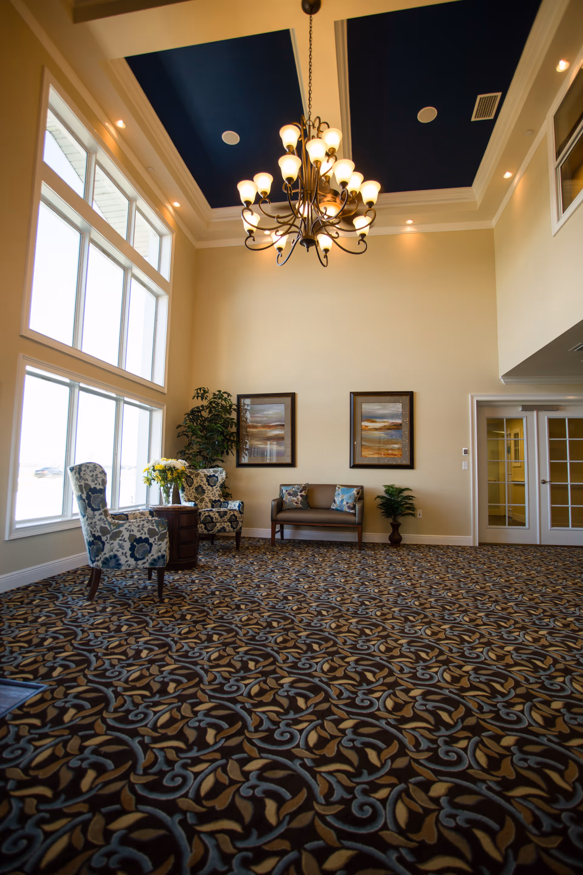 A spacious, well-lit sitting area with large windows on the left side allowing natural light to fill the room. The room features a patterned carpet with blue and brown tones, two floral upholstered armchairs with a small round wooden table between them holding a vase of yellow flowers, a small brown loveseat with decorative pillows, two framed landscape paintings on the wall, and two potted plants. A large chandelier with multiple lights hangs from the ceiling, which has a dark blue inset design.