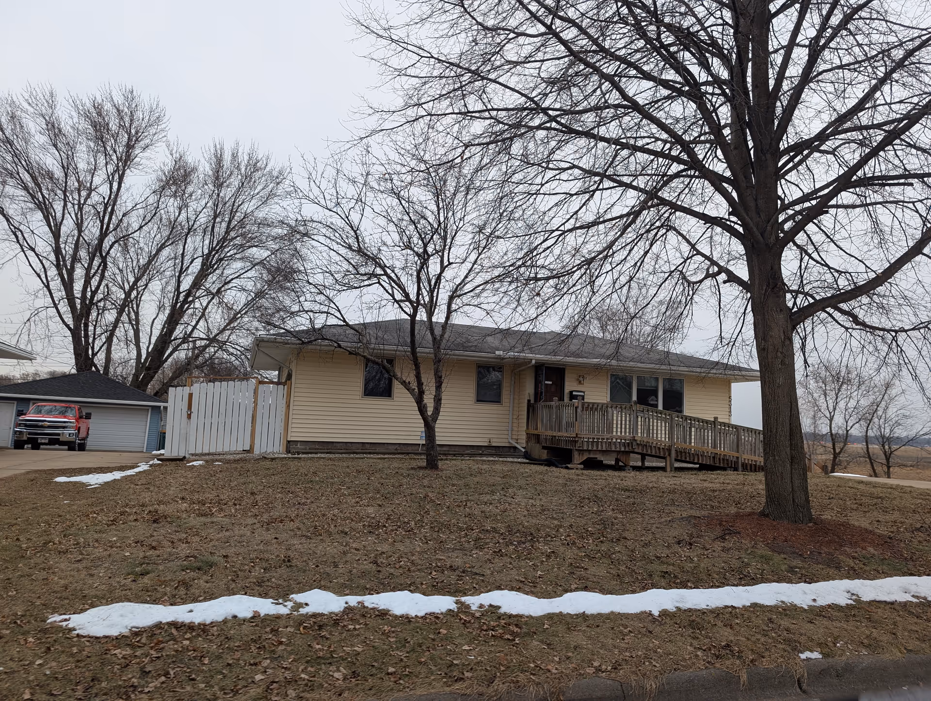 Single-story beige building with a wheelchair accessible wooden ramp leading to the front door. The building is surrounded by leafless trees and patches of snow on the grass. A red pickup truck is parked near a detached garage to the left.