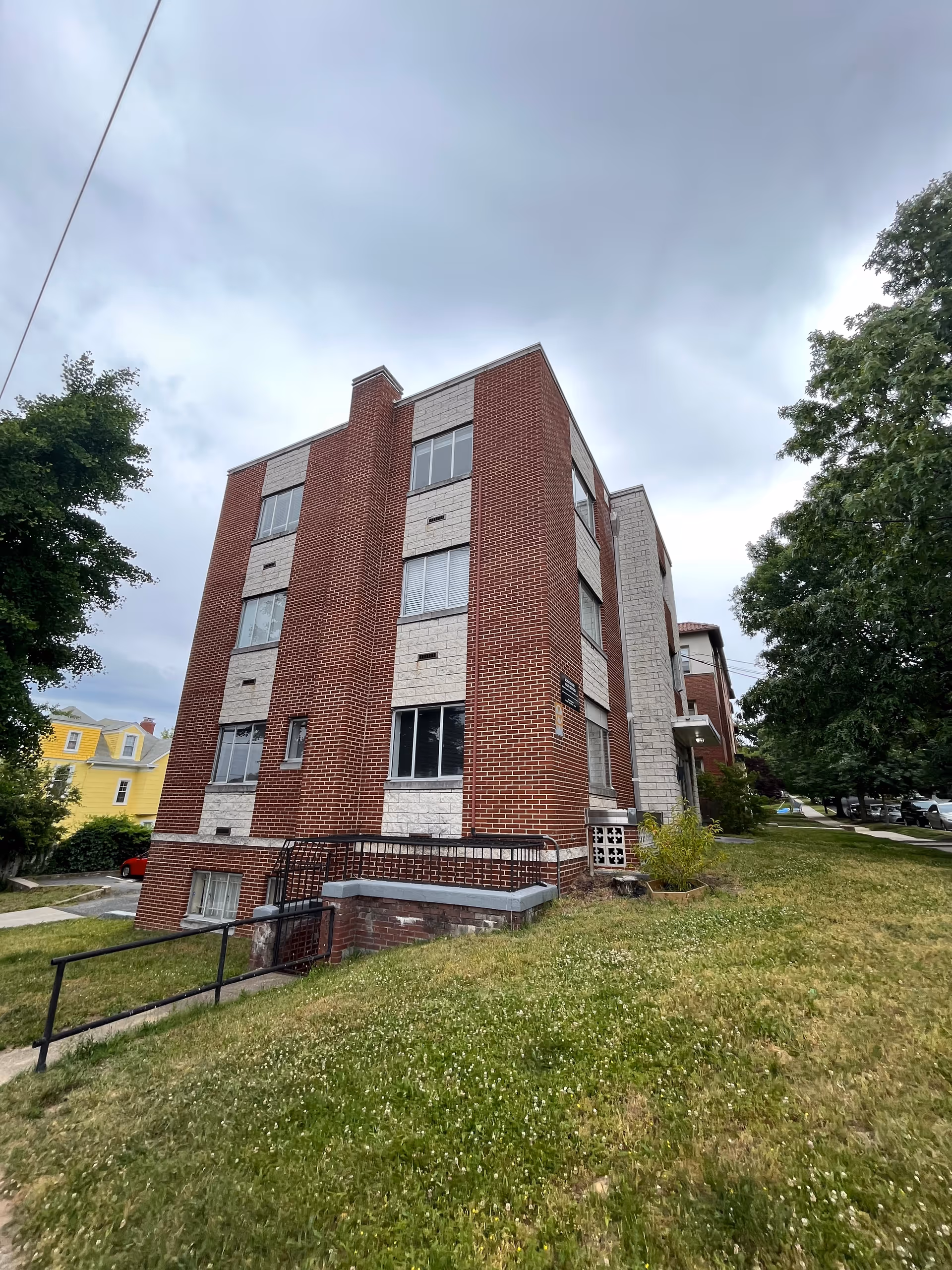 Three-story brick building with windows, a grassy lawn, and trees along a sidewalk.