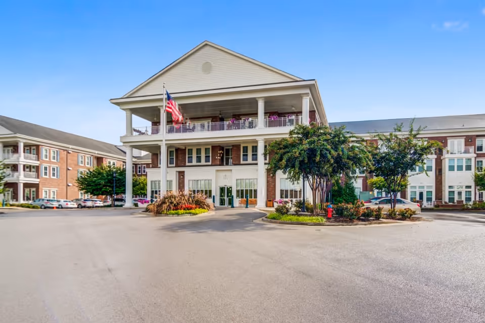 Front exterior of a multi-story senior living building with columns, balconies, an American flag, and a circular driveway.