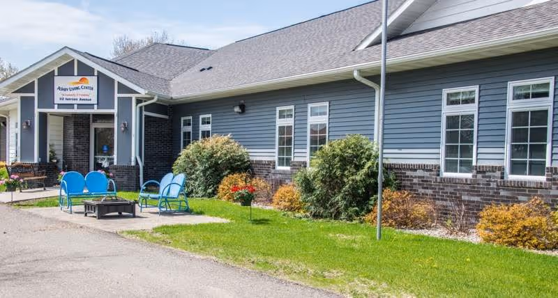 Front exterior of Ashby Living Center with entrance, blue siding, windows, landscaping, and outdoor seating.