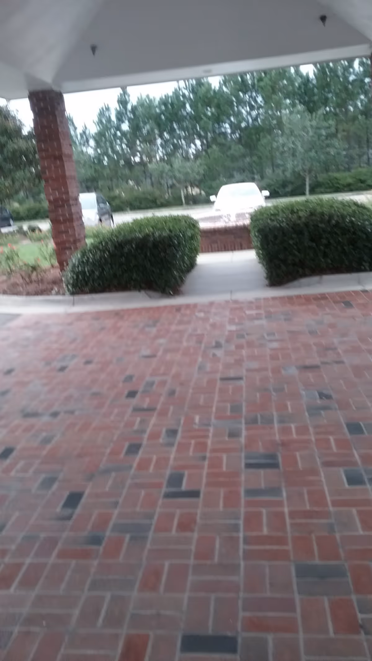 Brick-paved walkway toward an entrance, with trimmed hedges and parked vehicles nearby.