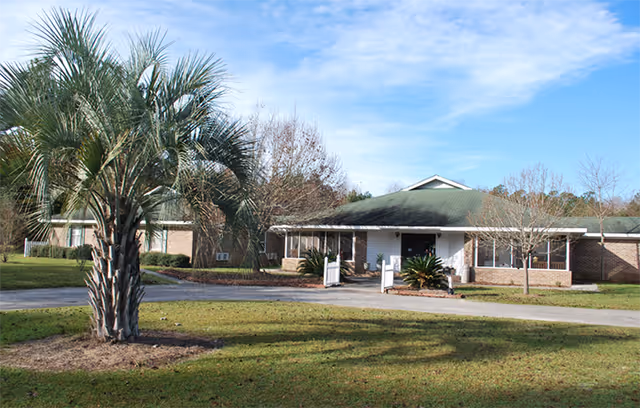Single-story assisted living facility exterior with a green roof, palm tree, and manicured lawn.