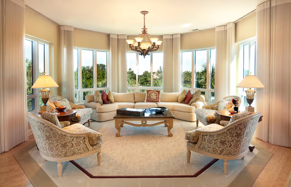 A bright and elegant living room with large windows offering a view of greenery outside. The room features a curved beige sectional sofa with decorative pillows, four patterned armchairs arranged around a wooden coffee table, two side tables with lamps, and a chandelier hanging from the ceiling.