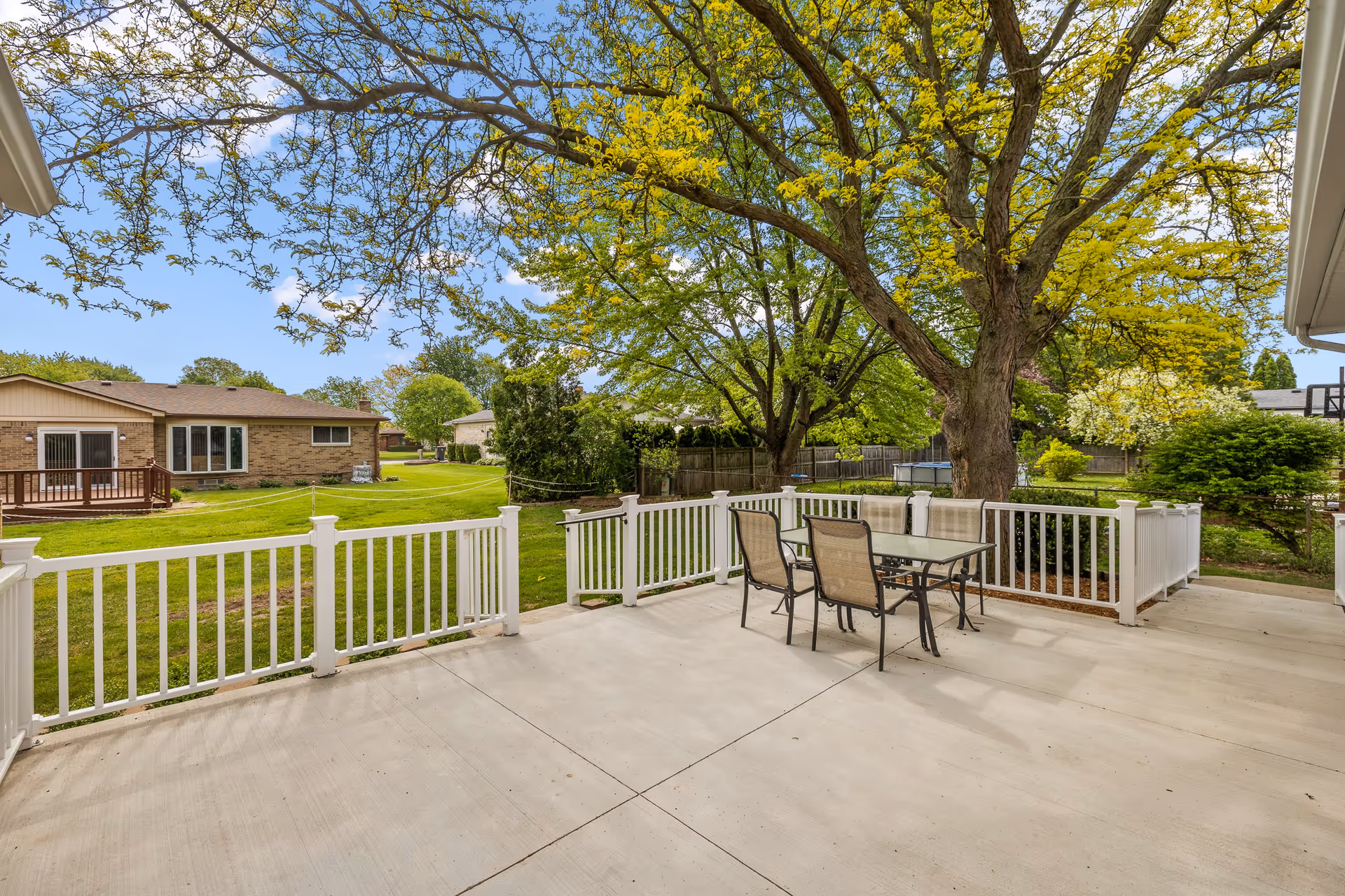 Outdoor patio area with a glass-top table and four chairs on a concrete surface, surrounded by white railing. Large trees with green and yellow leaves provide shade, and neighboring houses with green lawns are visible in the background under a partly cloudy sky.