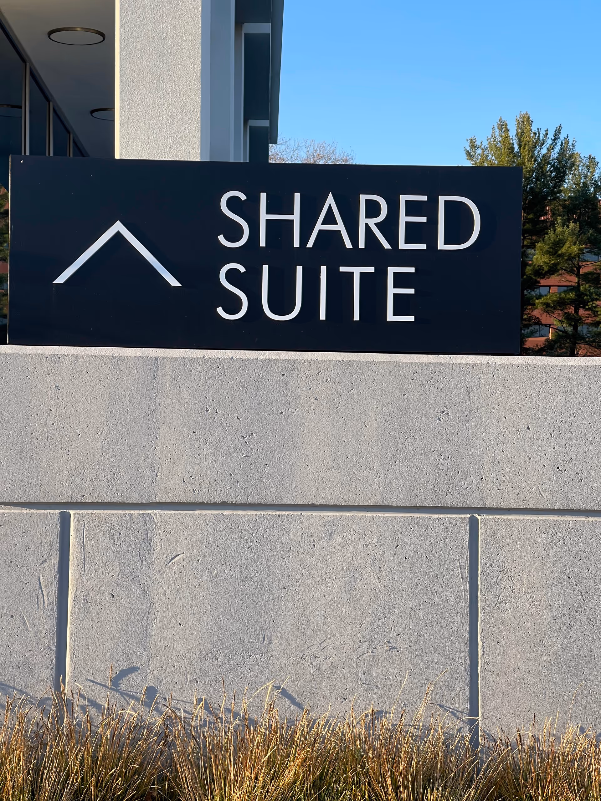 Dark sign reading SHARED SUITE on a concrete wall outside a modern building.