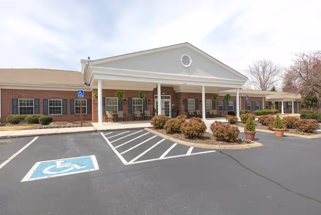 Front exterior view of a single-story brick building with white columns and a covered entrance. There are several windows with black shutters, potted plants, and bushes in front of the building. The parking lot includes a marked handicapped parking space.