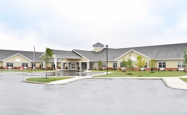 Low single-story senior living facility building with a covered main entrance, driveway, flagpole, and small landscaped lawn on a cloudy day.