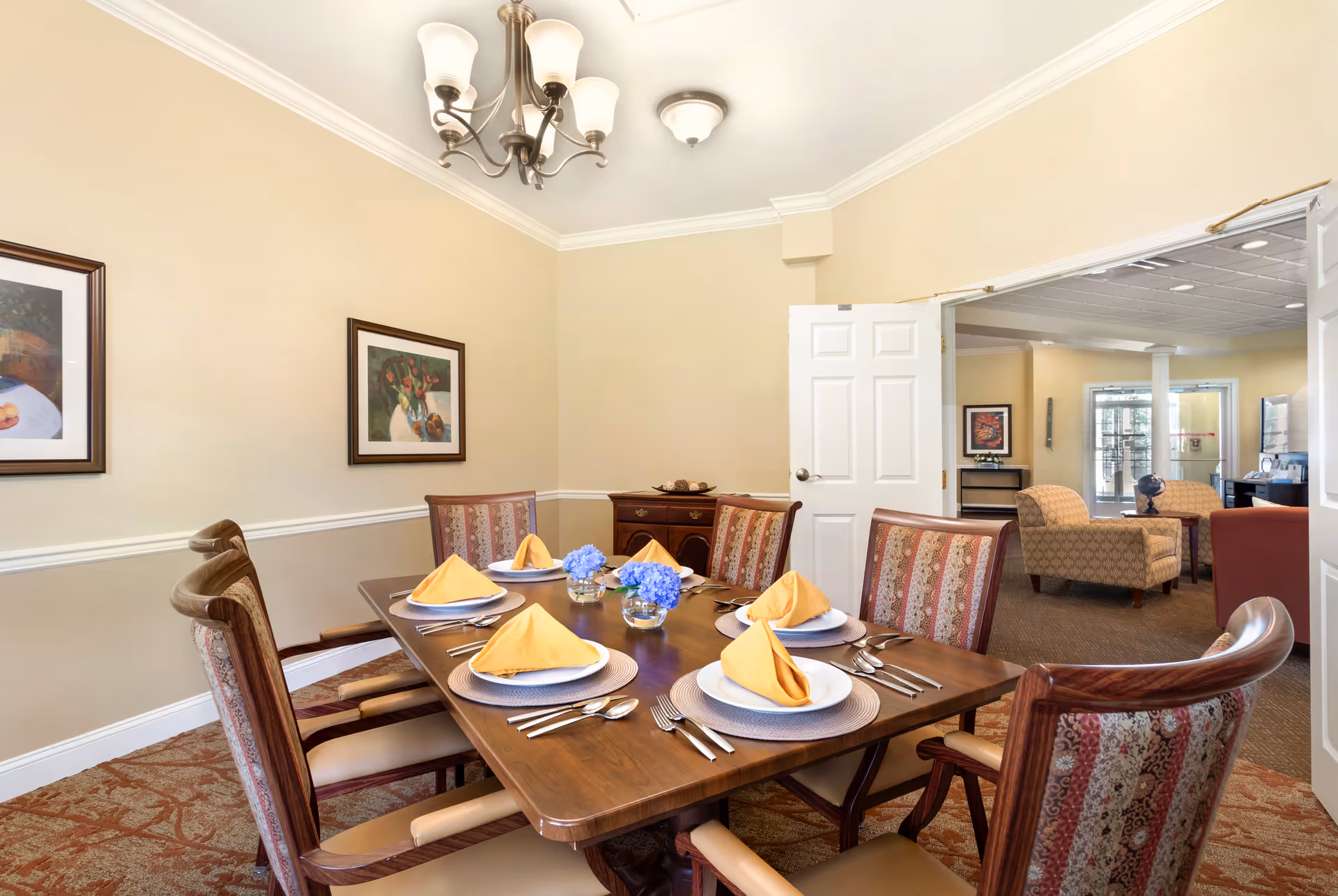 A dining room featuring a wooden table set with plates, silverware, and folded napkins. The room has light-colored walls, framed artwork, and a doorway leading to a lounge area with comfortable seating.