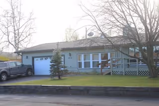 Single-story residential building with a light blue exterior, a white garage door, a small tree in the front yard, and a pickup truck parked in the driveway. There is a wooden ramp leading to the front entrance and leafless trees around the property.