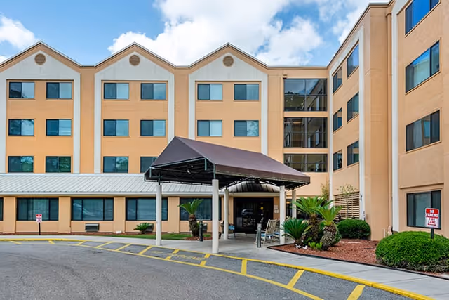 Exterior view of a multi-story senior living facility building with beige and light brown walls, multiple windows, and a covered entrance with a brown canopy. There are landscaped bushes and small palm trees near the entrance, and a driveway with yellow painted lines in front.