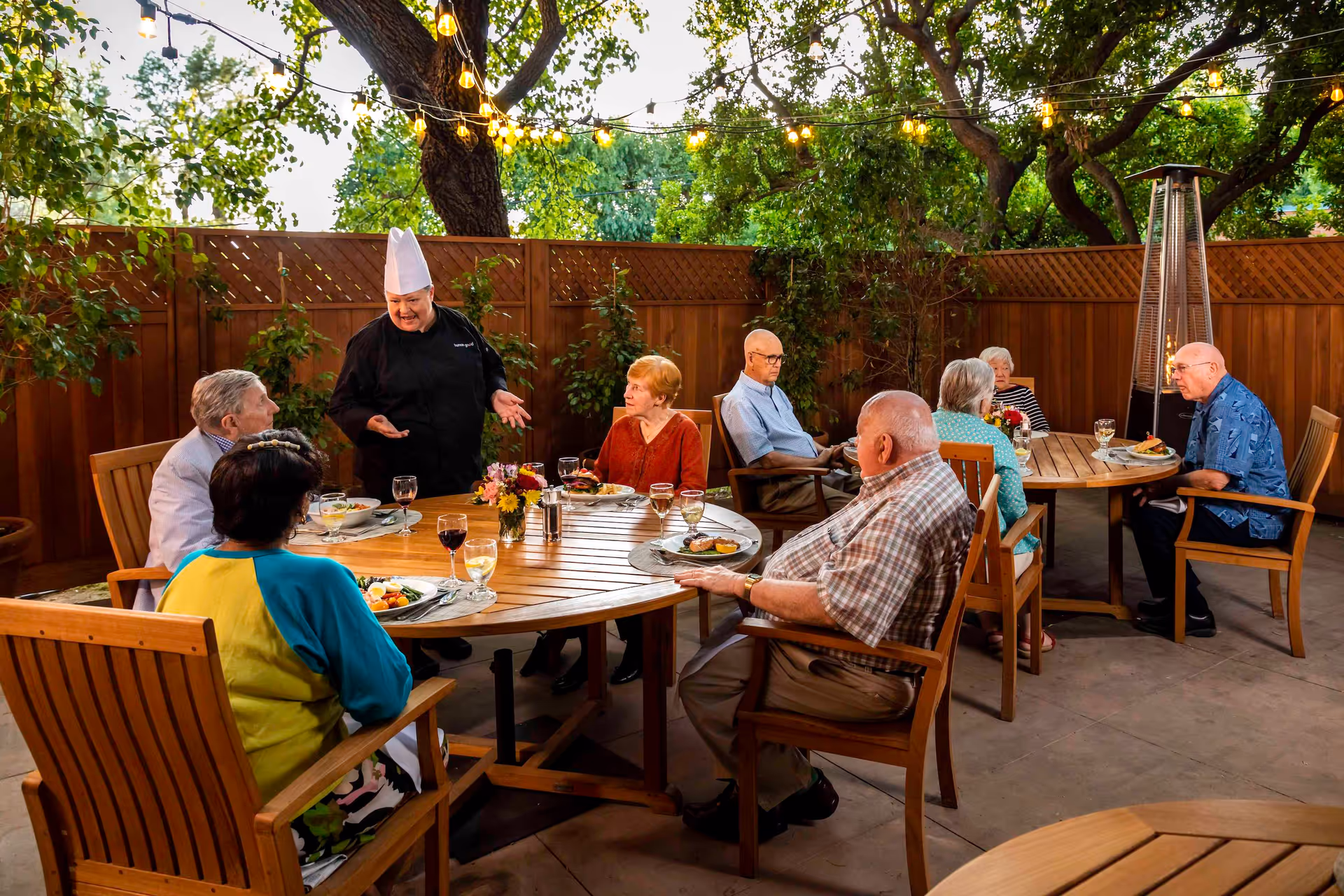 A group of elderly residents sit around wooden outdoor tables under string lights while a chef talks to them during an evening meal.