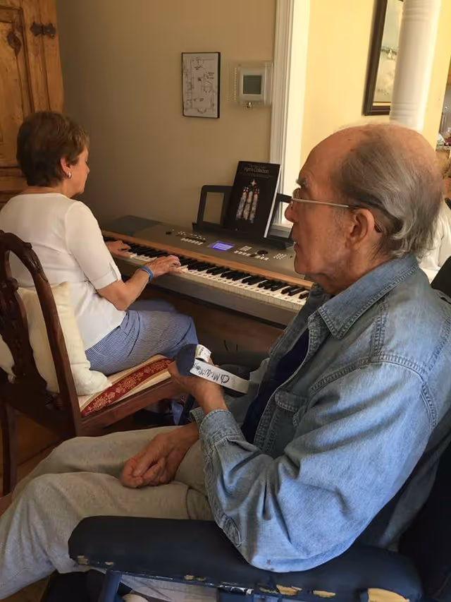 An elderly man sitting in a chair holding a small object, while an elderly woman sits at an electronic keyboard playing music in a cozy room with wooden furniture and framed pictures on the wall.
