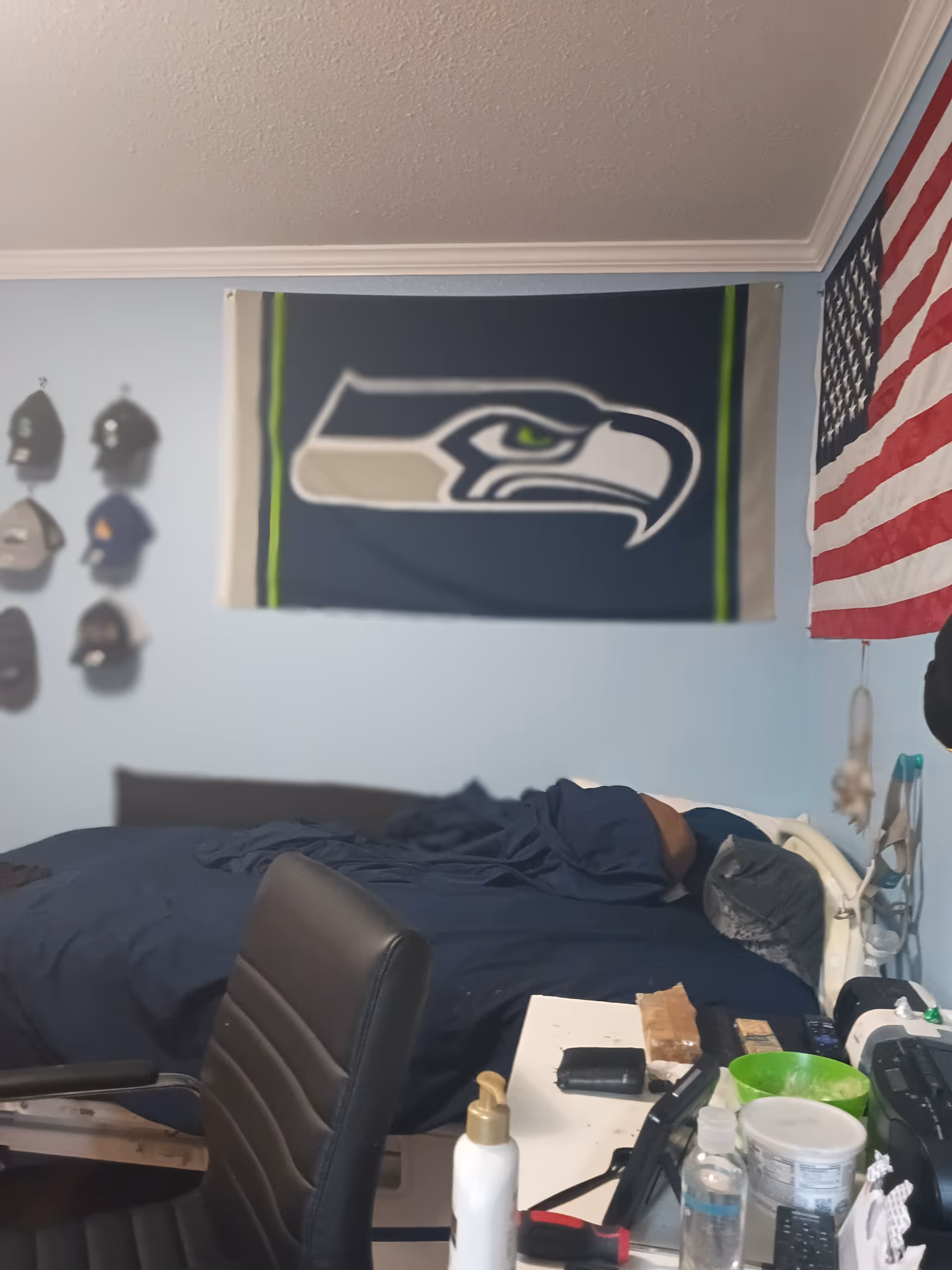Blue bedroom with bed, Seahawks banner, American flag, and cluttered desk.