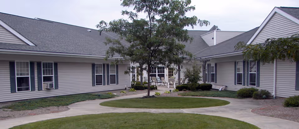 Courtyard of a single-story senior living community with a central tree, circular walkways, lawn and surrounding beige buildings with windows and patio chairs.