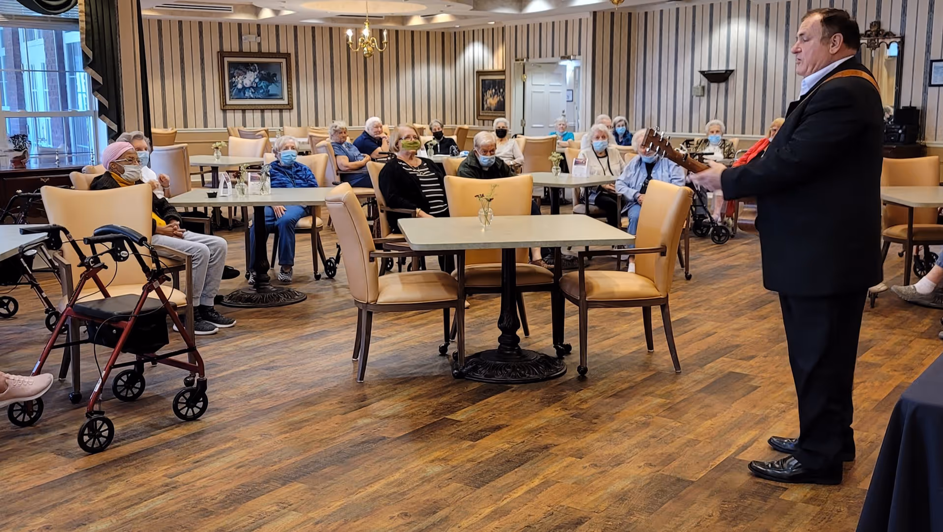 A group of elderly people seated at tables in a common room, attentively watching a man playing a guitar. The room has wooden flooring, beige chairs, and striped wallpaper. Some attendees are wearing face masks.