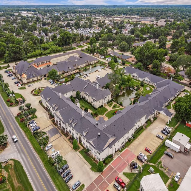 Aerial view of Montclair Park Assisted Living facility showing multiple connected buildings arranged around a landscaped courtyard with a pond and walking paths. Surrounding the facility are parking areas, roads, and a suburban neighborhood with many trees and houses.