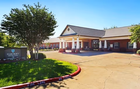Exterior view of a senior living facility building with a covered entrance, surrounded by trees and a driveway with a red curb marked 'FIRE LANE NO PARKING'. A sign near the entrance reads 'ELMCROFT SENIOR LIVING'.