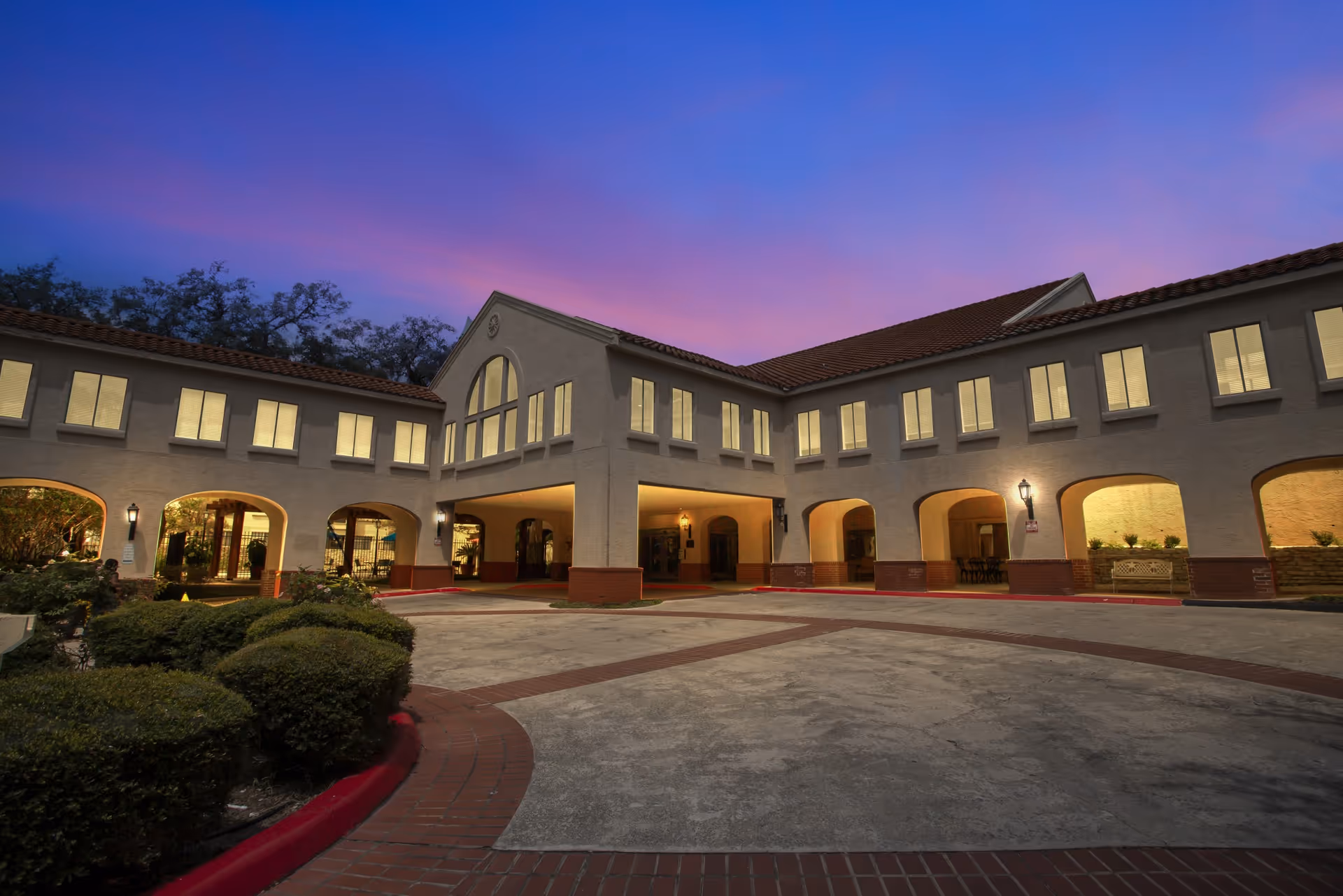 Exterior view of a two-story building at dusk with warm lights glowing from the windows. The building has an arched entrance and a tiled roof, surrounded by bushes and a paved driveway with red brick accents.