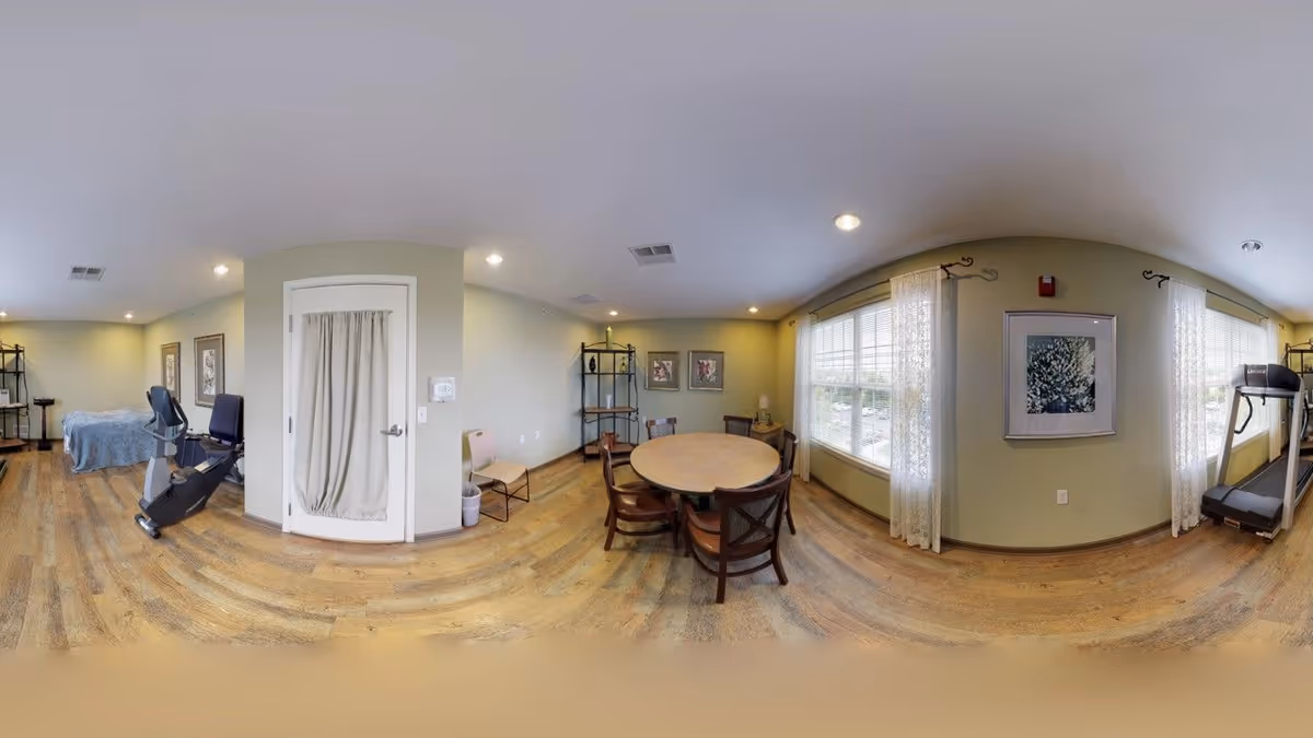 Panoramic view of a cozy communal area with a round table, windows, and exercise gear.