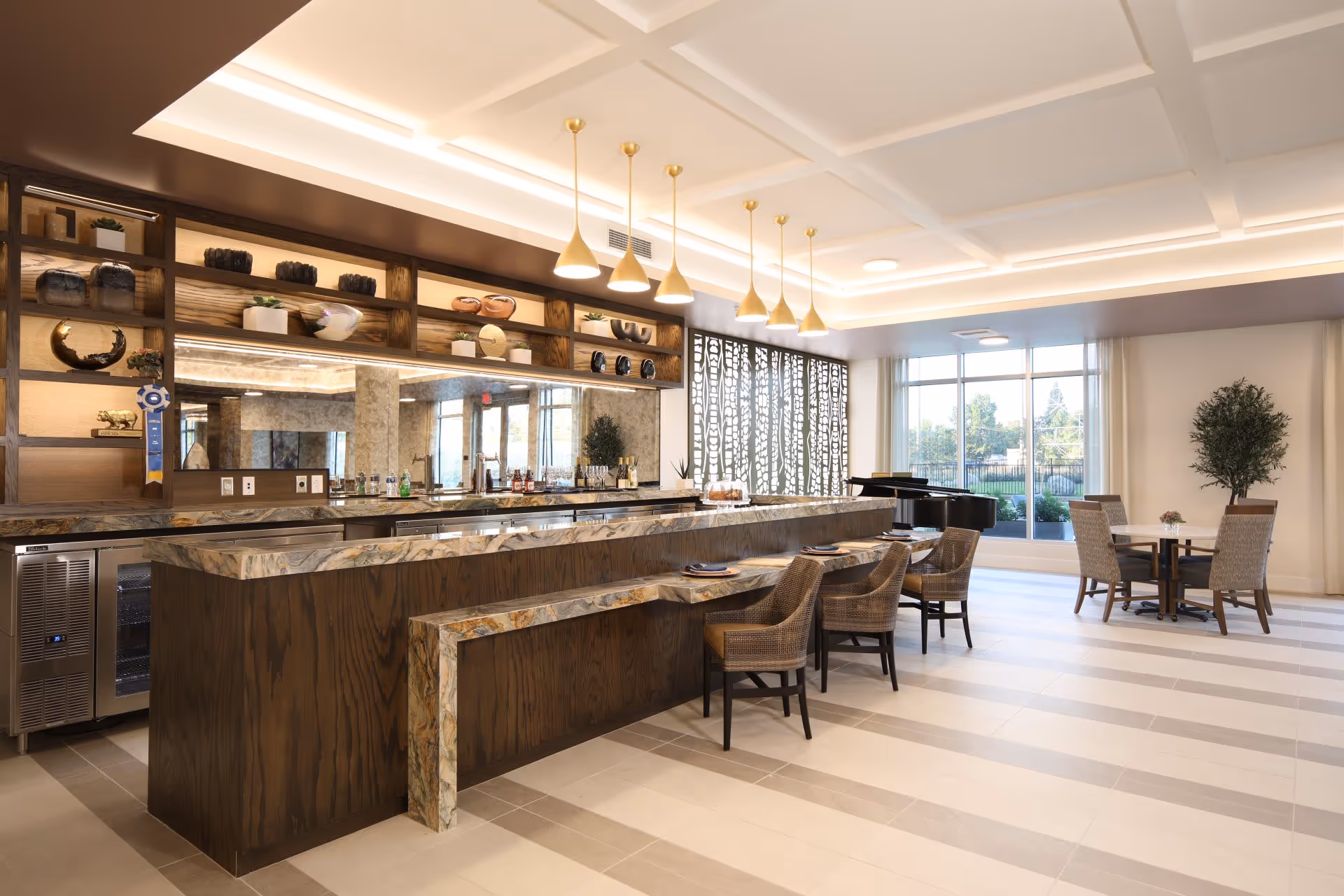 A modern and spacious interior area featuring a long marble countertop bar with six wicker chairs. Above the bar, there are six gold pendant lights hanging from a white coffered ceiling. Behind the bar, shelves display decorative items and a large mirror reflects the room. To the right, there is a round table with four chairs near large windows that let in natural light, and a black grand piano is visible in the background.