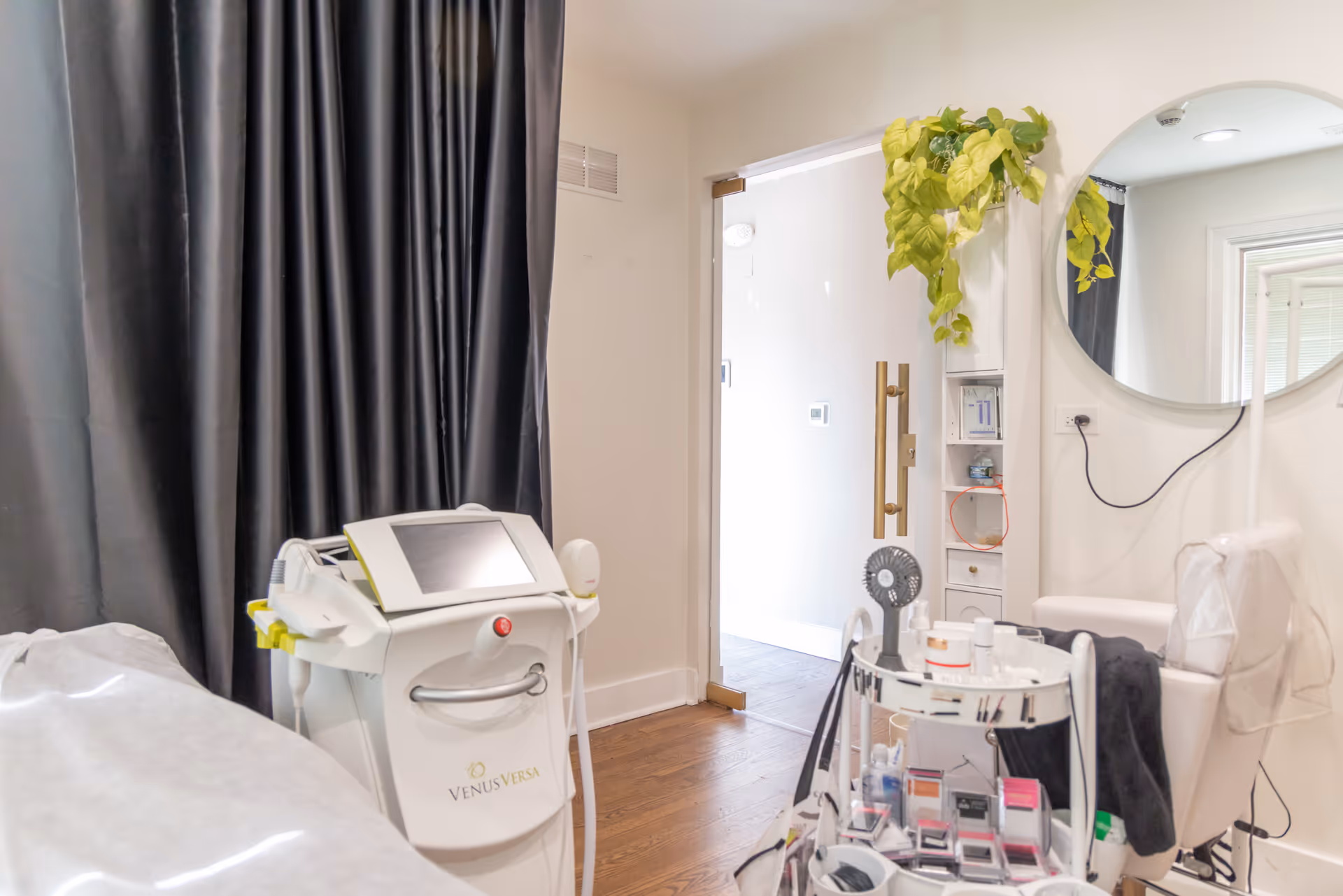Interior medical treatment room with Venus Versa device, cart, curtain, mirror, and plant.