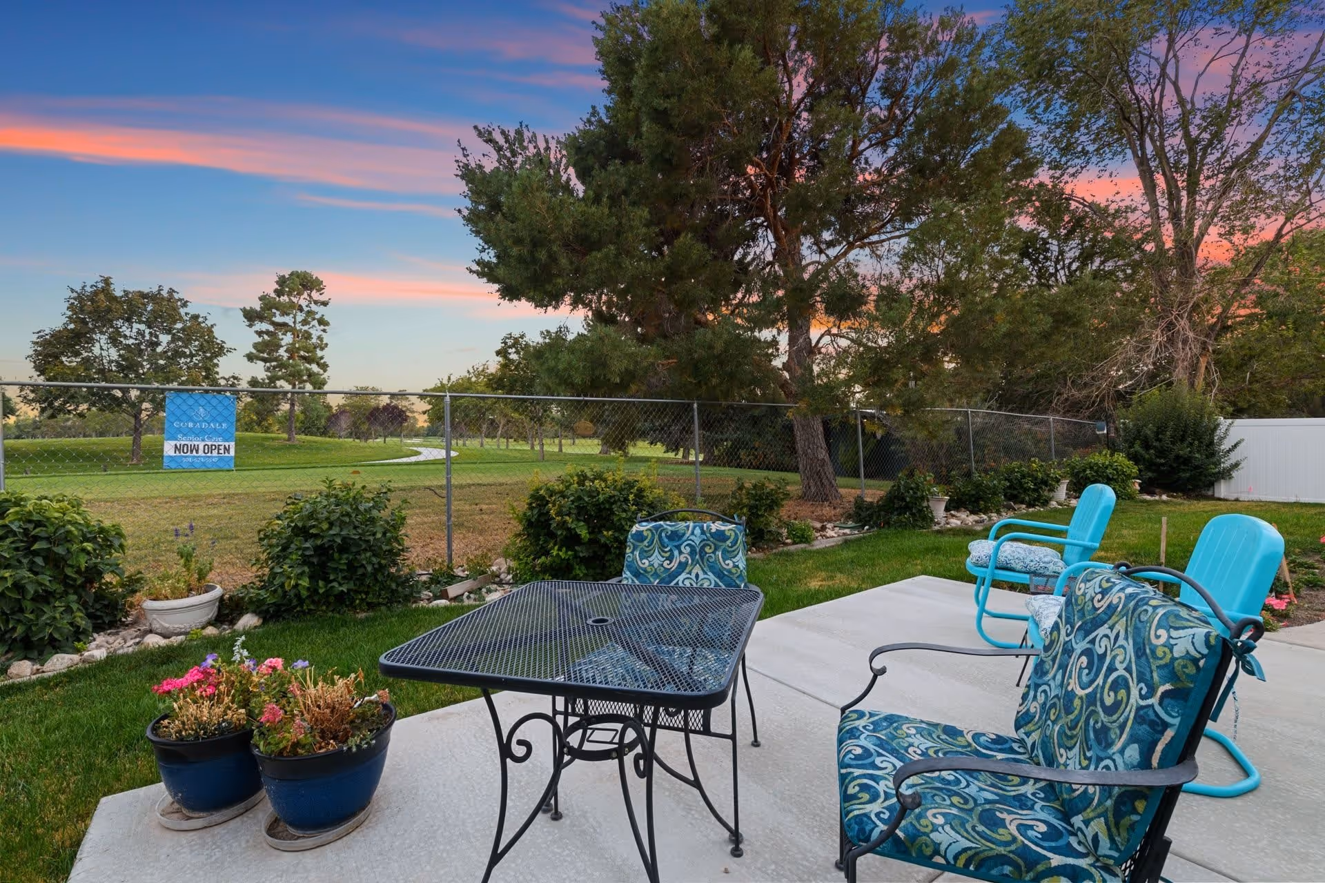 Patio with a metal table, patterned and turquoise chairs, and potted plants overlooking a fenced grassy yard and trees under a colorful sunset sky.
